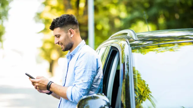 A man in a white shirt looking at his phone while standing next to a car