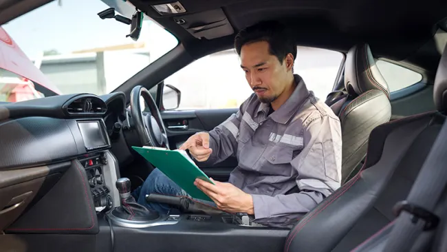An auto mechanic holding a clipboard while seated in the driver's seat 