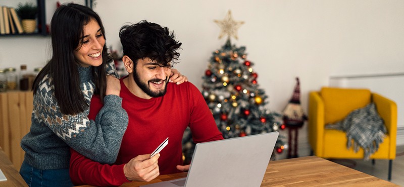 Man and woman looking at a laptop, holding a credit card, with a Christmas tree in the background