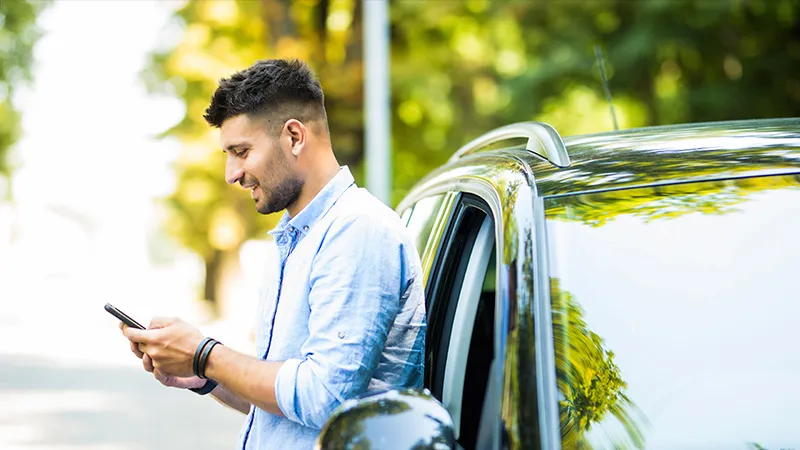 A man in a white shirt looking at his phone while standing next to a car