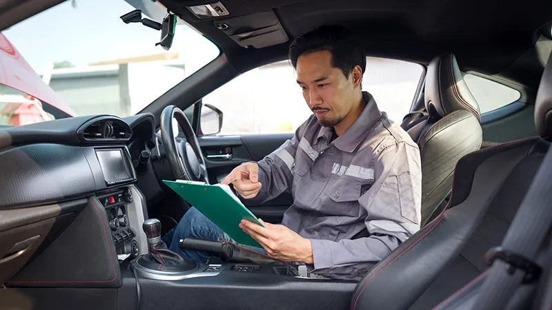 An auto mechanic holding a clipboard while seated in the driver's seat 