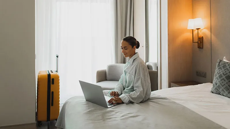 A woman who is seemingly working on a laptop while sitting on the edge of a hotel bed